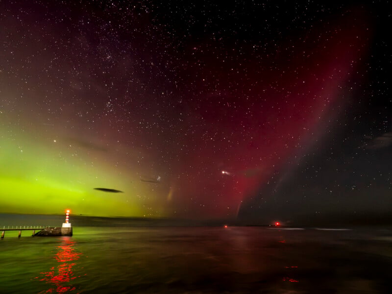 A vibrant night sky shows green and red auroras above a calm sea, with stars visible. Two lit beacons mark the end of a pier and a distant point on the water, reflecting their lights on the waves.