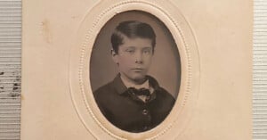 A sepia-toned vintage photograph of a young boy with short dark hair, wearing a suit jacket and bow tie, set in an oval-shaped frame with a decorative border on a cream-colored background.