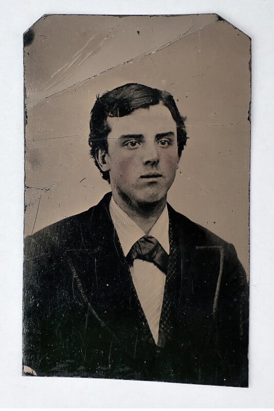 A vintage black and white portrait of a young man with dark hair, wearing a suit and bow tie, looking slightly to the side. The photo has scratches and worn edges, reflecting its age.