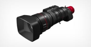 A professional Canon video camera with a large, detachable zoom lens and a wide, square lens hood, shown on a white background. The lens features ridged focus and zoom rings with a red accent near the base.