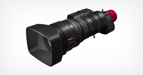 A professional Canon video camera with a large, detachable zoom lens and a wide, square lens hood, shown on a white background. The lens features ridged focus and zoom rings with a red accent near the base.