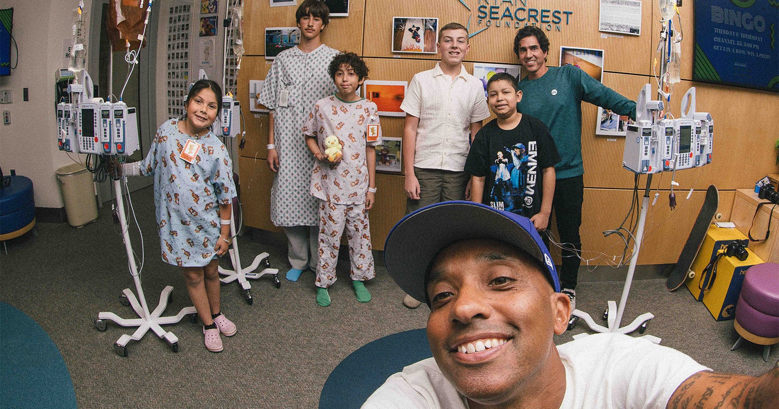 A group of children in hospital gowns and adults pose together and smile in a hospital room, surrounded by medical equipment. One man is taking a selfie in the foreground.