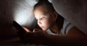 A young girl lies under a blanket in the dark, illuminated by the light from a smartphone she is holding, focused intently on the screen.