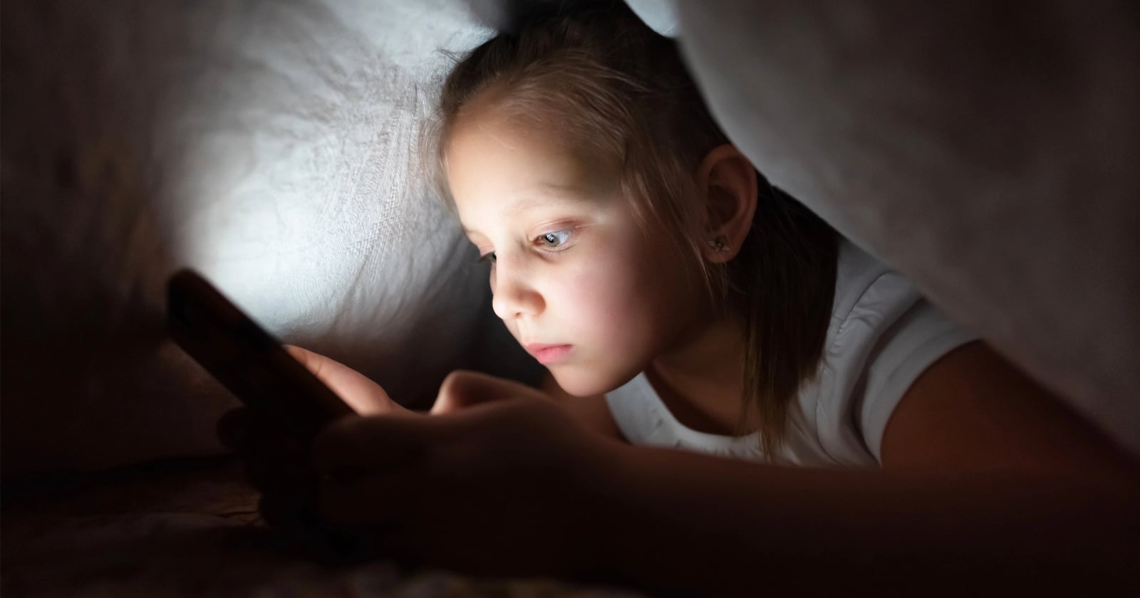 A young girl lies under a blanket in the dark, illuminated by the light from a smartphone she is holding, focused intently on the screen.