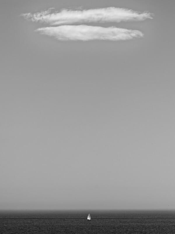 A black and white photo of a lone sailboat on the horizon beneath a vast sky, with a single cloud near the top of the image, creating a minimalist and serene seascape.