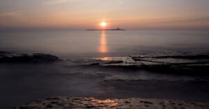 A calm sea at sunset with the sun low on the horizon, casting a golden reflection on the water. Rocky shoreline in the foreground and a small island with a lighthouse in the distance.