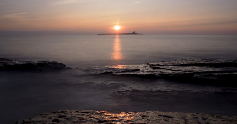A calm sea at sunset with the sun low on the horizon, casting a golden reflection on the water. Rocky shoreline in the foreground and a small island with a lighthouse in the distance.