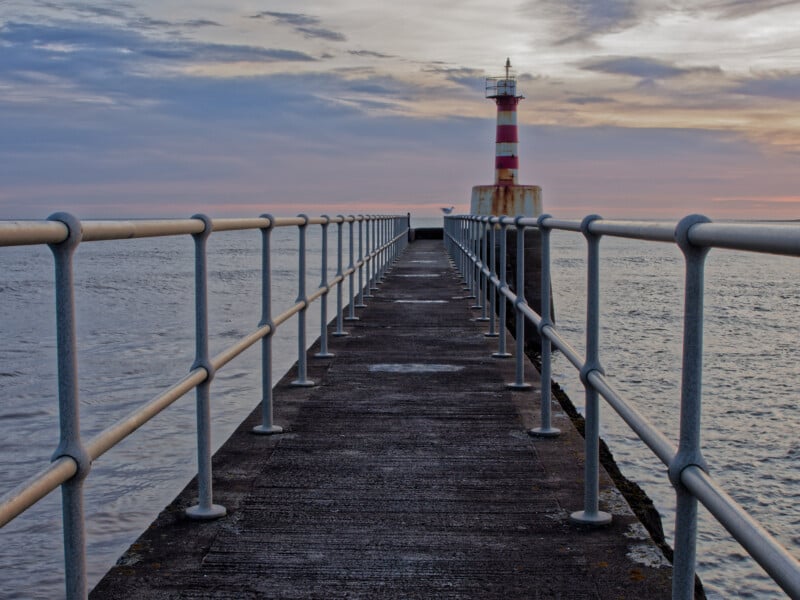 A concrete pier with metal railings leads to a red-and-white striped lighthouse at the end, surrounded by calm sea under a cloudy sunset sky.