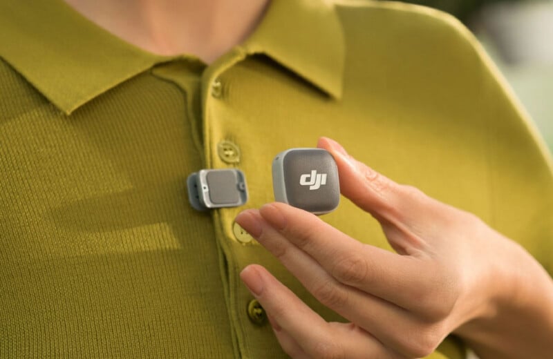A person in a green shirt clips a small, square DJI wireless microphone to their collar, while holding another similar device in their hand.