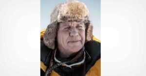 An older person wearing a heavy fur-lined winter hat and a yellow insulated jacket stands outside in bright sunlight, with a camera strap around their neck.