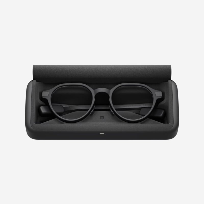 A pair of black smart glasses with round lenses resting in an open, black rectangular case against a plain white background.
