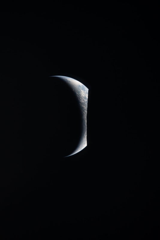 A crescent-shaped view of Earth is visible against a black background, with most of the planet in shadow and only a thin, sunlit edge showing clouds and landforms.