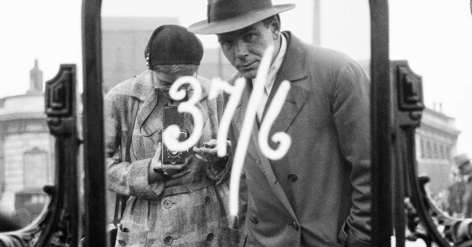 A black and white photo shows a woman with a camera and a man in a trench coat and hat, reflected in a mirror with "37%" written on it, in an urban setting.