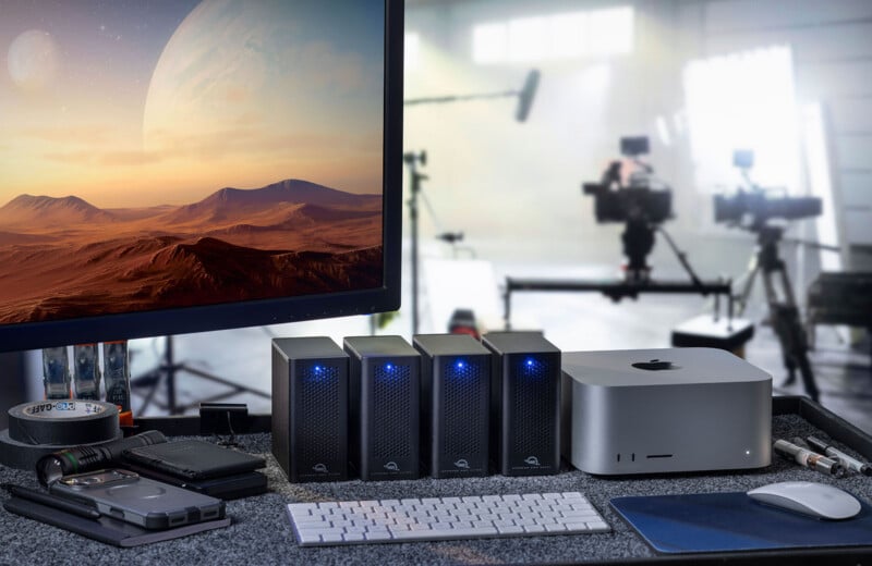 A modern editing desk with a computer monitor showing a planetary landscape, several external hard drives with blue lights, a Mac Studio, a white keyboard, and film equipment in a blurred background.