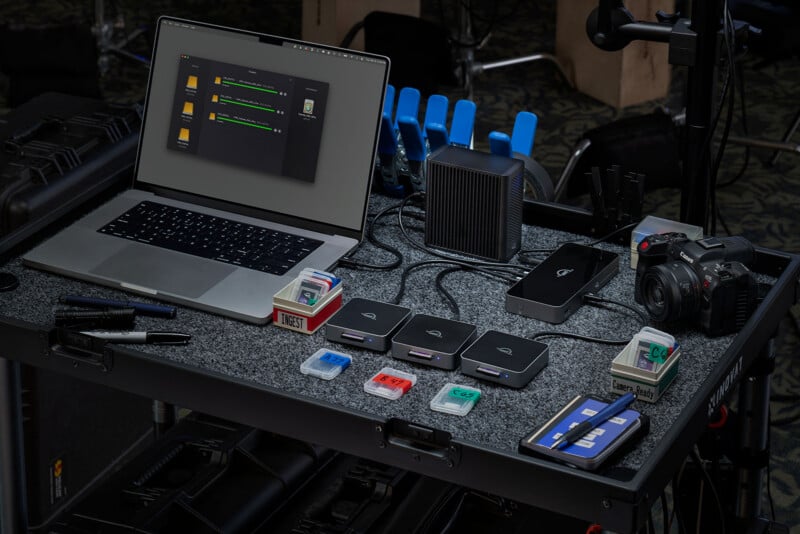 A laptop on a rolling cart is connected to card readers, memory cards, a camera, and various storage devices, with cables and other equipment organized neatly on the gray surface.