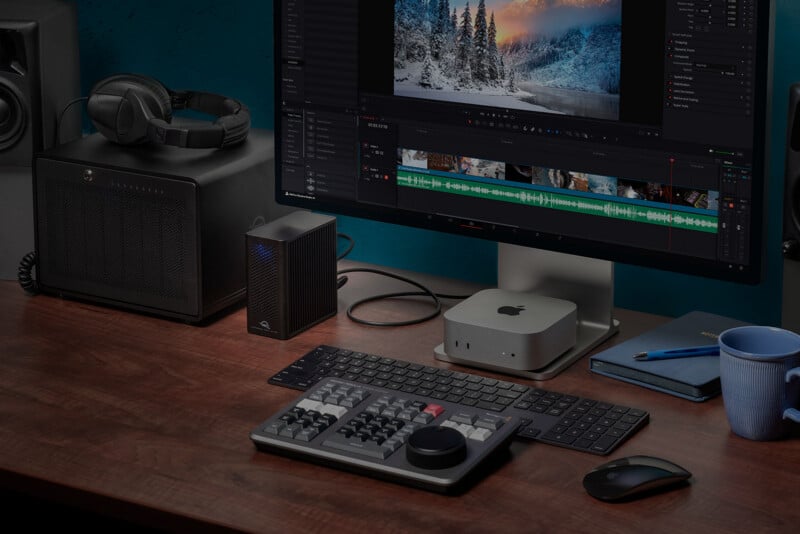 A desk setup with a computer monitor displaying video editing software, a keyboard, a control panel, a mouse, a Mac Mini, headphones, speakers, an external drive, and a blue mug.
