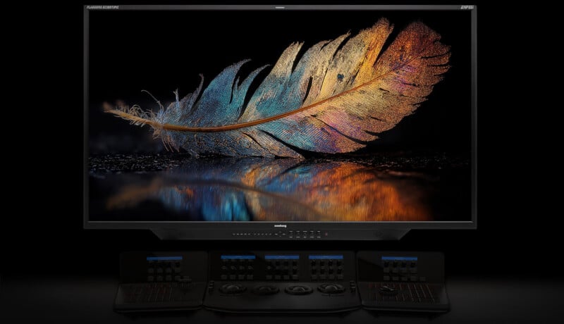 A large monitor displays a detailed, colorful feather with gold, blue, and orange hues, reflected on a shiny surface. In front of the monitor are multiple panels with dials and buttons, likely for video editing or color grading.