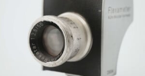 Close-up of the lens on a vintage Flexameter camera, showing engraved text "f/2.8 50 Dewatar" on the lens ring, with a metallic, slightly worn appearance and part of the camera body visible in the background.
