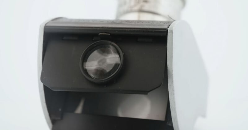Close-up of a metal and plastic component with a circular glass lens in the center, possibly part of a camera or optical device, against a plain white background.