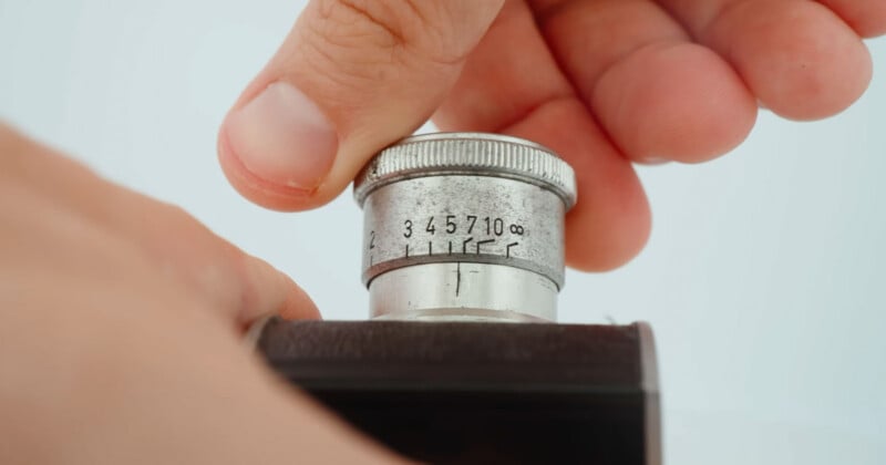 A close-up of a hand adjusting the focus ring on a vintage camera lens, showing engraved numbers and distance markings from 1 to infinity.