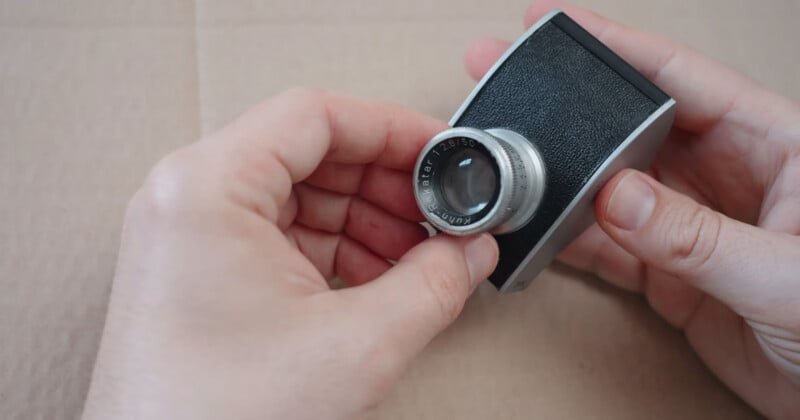 A person holds a small vintage camera with a textured black grip and a silver lens against a beige background.