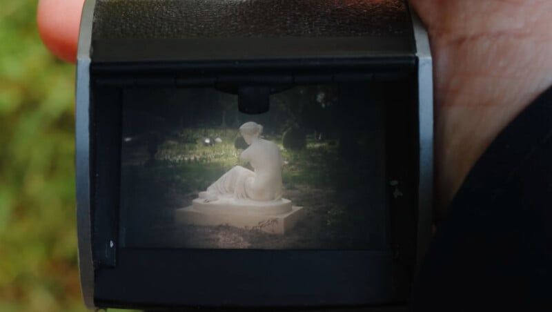 A close-up view through a camera’s viewfinder shows a white statue of a seated woman on a pedestal in an outdoor, green setting. A person's hand is holding the camera.