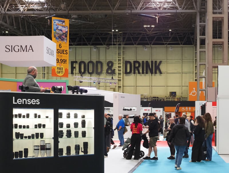 People attend a photography trade show with booths displaying camera lenses and gear. Large "FOOD & DRINK" signage is visible on the back wall, and various banners and displays fill the busy exhibition hall.