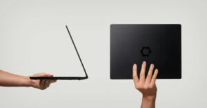 Two hands hold a slim black laptop: one hand supports it open from the side, while the other holds it closed from the back, showing a flower-shaped logo on the cover against a plain background.