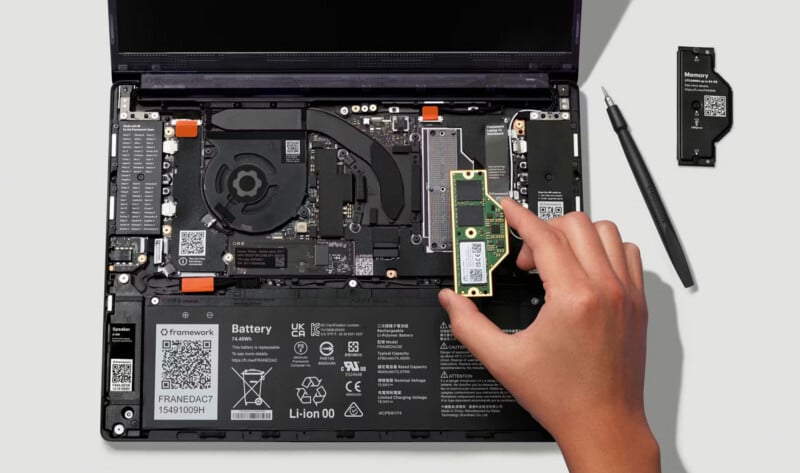 A hand is installing a RAM module into a laptop with its back cover removed, showing internal components like the battery, fan, and motherboard. A screwdriver and another memory module are beside the laptop.