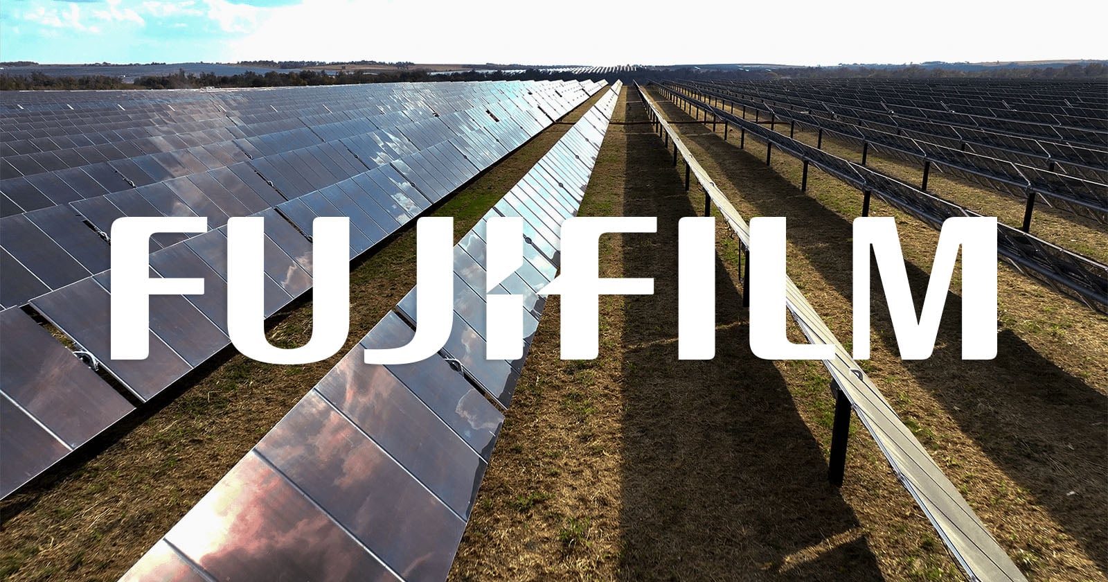 Rows of solar panels in a large field under a bright sky, with the word "FUJIFILM" in bold white letters overlaid across the center of the image.