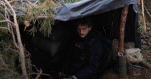 A person sits inside a makeshift shelter made of branches and a tarp in a wooded area. The surroundings appear rugged, with bags and supplies placed nearby. The person looks down, partially hidden in shadow.