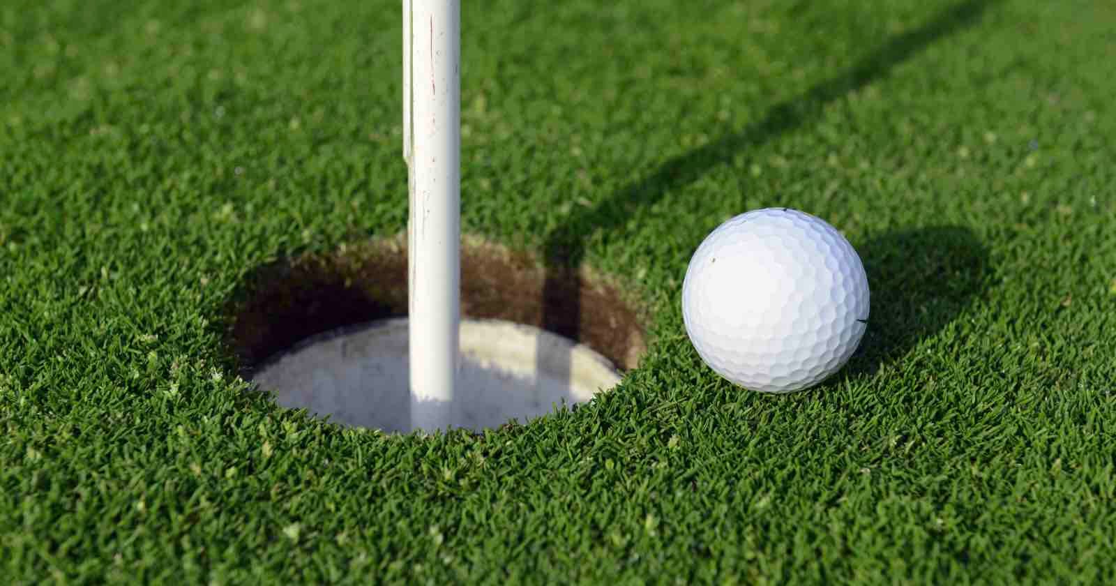 A close-up of a white golf ball resting on green grass next to the edge of a golf hole with the flagstick in place.