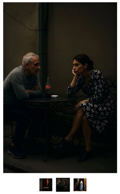 A man and a woman sit at a small round table outdoors, facing each other. The woman rests her chin on her hand, looking thoughtful, while the man leans forward. A Coca-Cola bottle and ashtray are on the table.