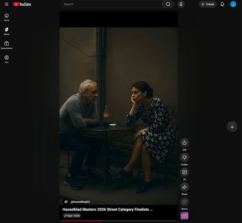 A man and woman sit across from each other at a small round table outdoors, both leaning in and engaged in conversation. There is a glass bottle on the table. The setting appears dimly lit, creating an intimate atmosphere.