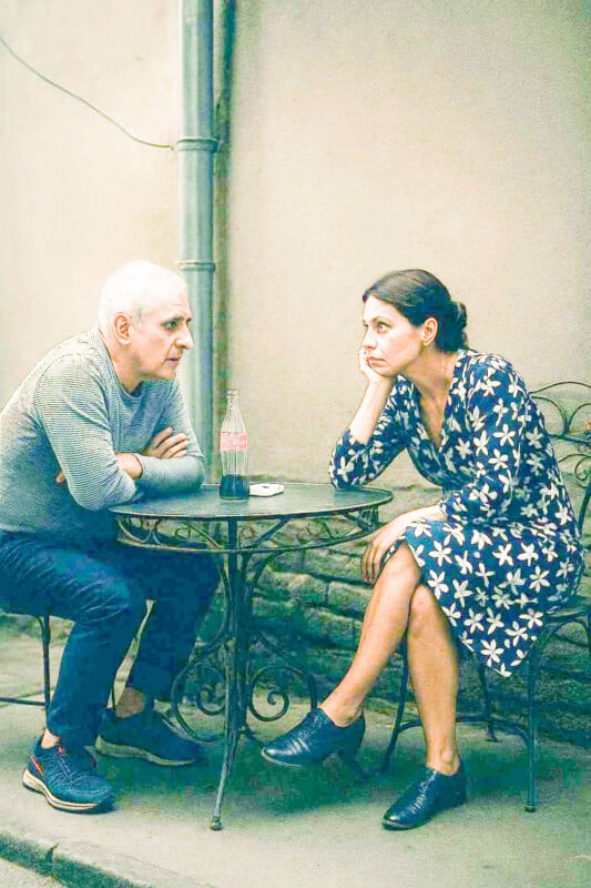 An older man and a younger woman sit at a small outdoor table with a drink between them. The man leans forward with arms crossed and the woman rests her head on her hand, looking thoughtful.