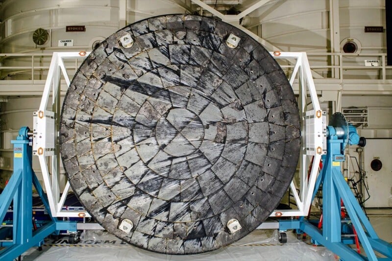 A large, circular heat shield with a patchwork of charred tiles is mounted vertically on a blue metal support structure inside an industrial facility.