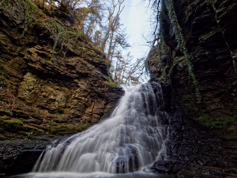 A waterfall cascades down layered, rocky cliffs surrounded by moss and trees, with water flowing smoothly over dark rocks under a bright sky.