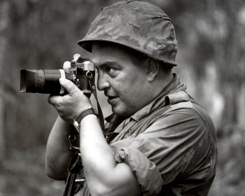 A man wearing a military helmet and uniform holds a camera up to his eye, appearing to take a photograph outdoors. The image is in black and white.