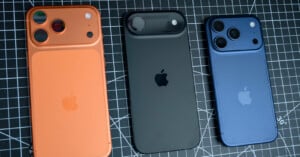 Three iPhone models in orange, black, and blue are displayed on a black grid surface. Each phone shows the Apple logo and has varying camera designs on the back.