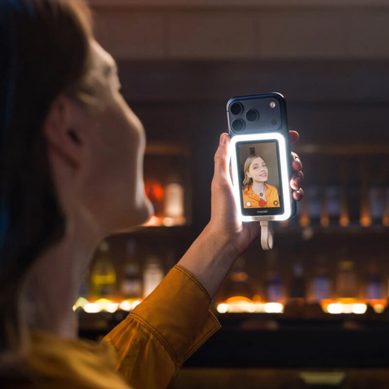 A person in an orange shirt holds a smartphone with a lit ring light attachment, taking a selfie in a dimly lit bar or restaurant. The phone screen shows their smiling reflection.