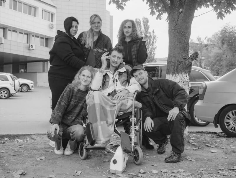 A group of six people, including a person in a wheelchair covered with a blanket, pose together outdoors near a tree and parked cars, smiling for the photo.