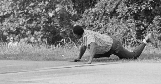 A man in a plaid shirt and dark pants crawls on hands and knees across a sidewalk, with grass and trees in the background. Two people sit in the distance on the left side of the image.