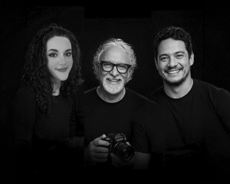 Three people pose against a black background. The woman on the left has curly hair, the man in the center has glasses and holds a camera, and the man on the right has short hair. All three are smiling and wear black shirts.