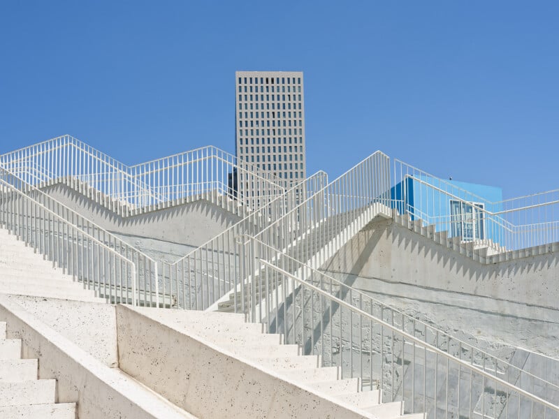 Staircases with white railings zigzag upward against a blue sky, with a tall, grid-patterned building in the background and modern blue structures visible on the right.