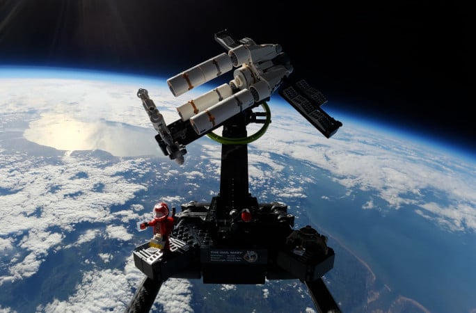 A LEGO model of a space shuttle and astronaut is photographed high above Earth, with clouds, land, and the planet’s curve visible against the blackness of space in the background.