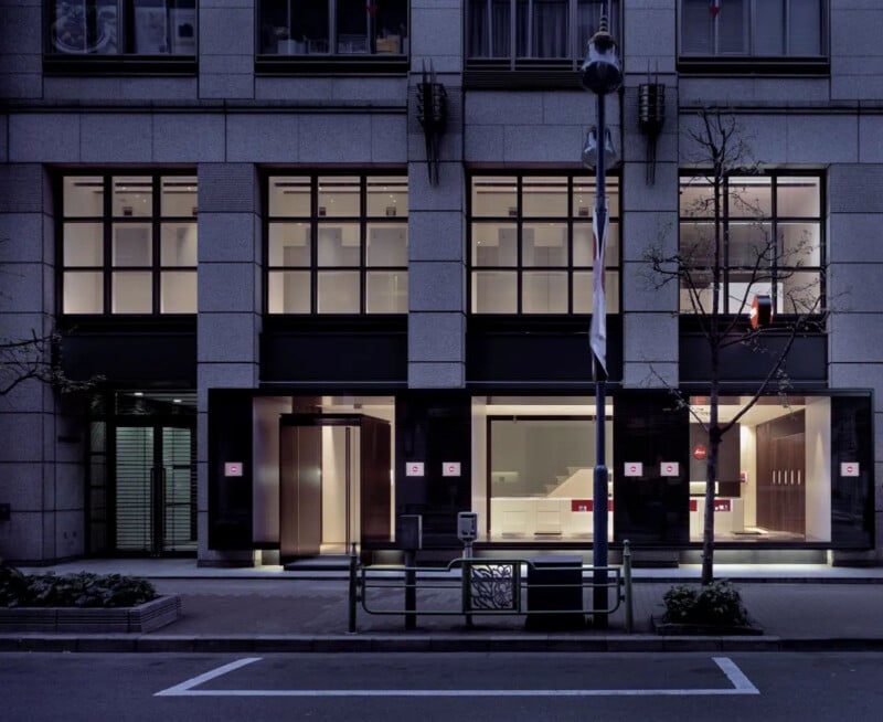 A modern office building at dusk with large windows, illuminated interior lighting, and a mostly empty sidewalk in front. Sparse trees and a metal bike rack are visible along the street.