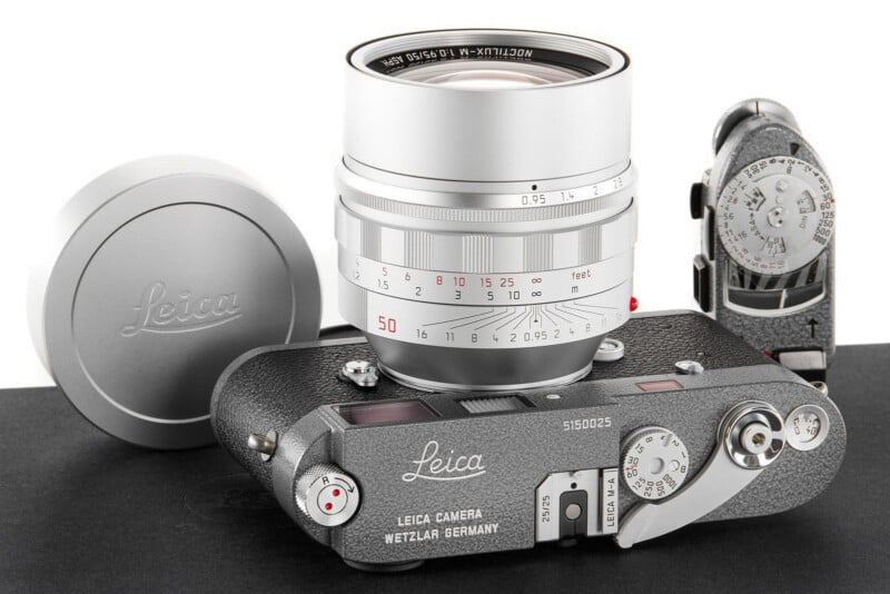 A silver Leica camera with a mounted lens sits on a black surface next to a Leica lens cap and a vintage light meter. The camera body displays the Leica logo and "LEICA CAMERA WETZLAR GERMANY.