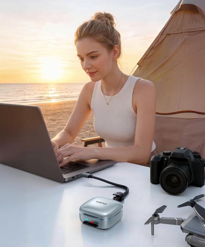 A woman works on a laptop at a beachside campsite during sunset, with a camera and a drone on the table beside her. A portable power bank is connected to the laptop.
