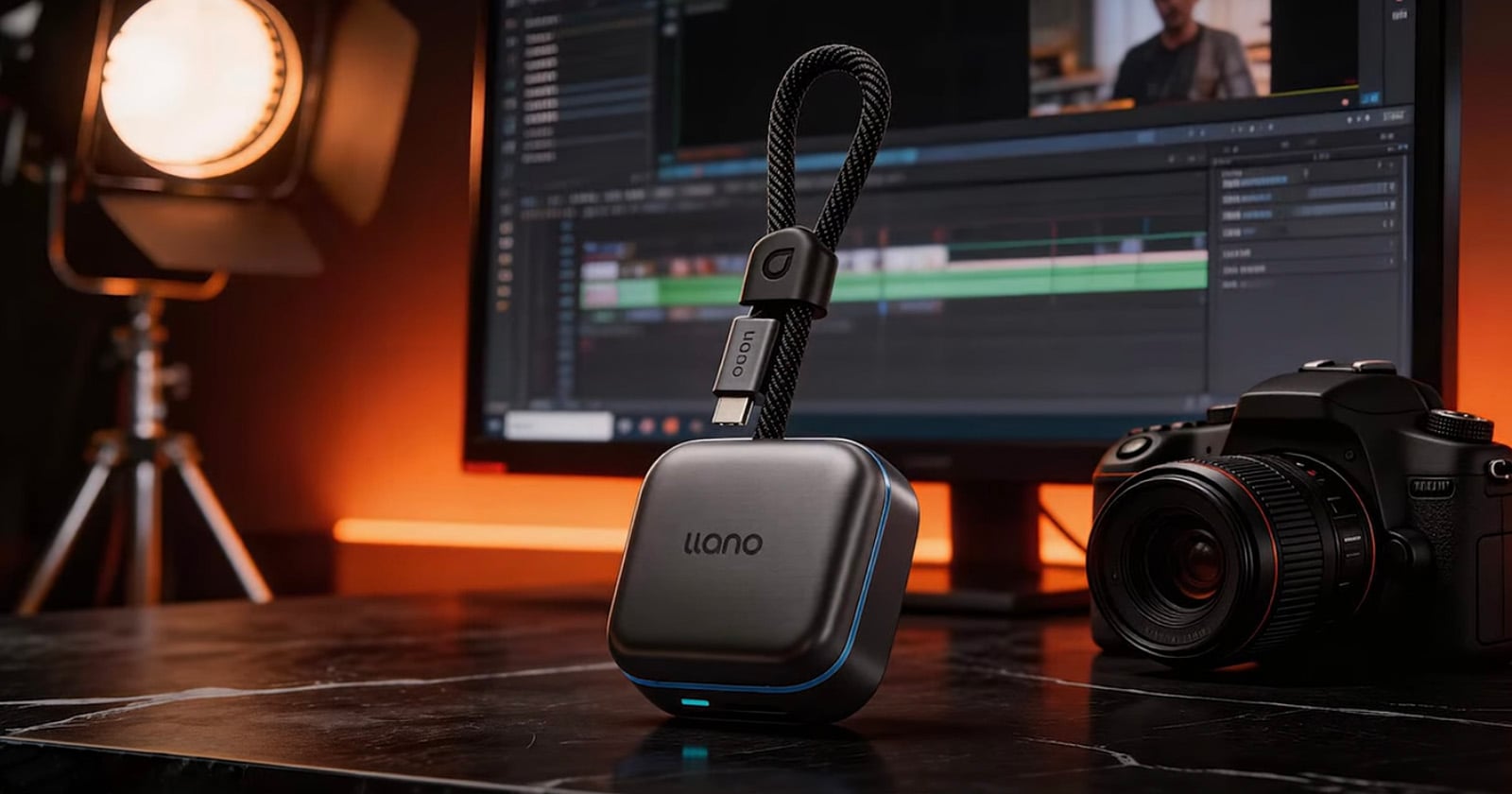 A llano portable device with a braided cable stands on a dark marble desk, in front of a camera, a computer monitor displaying a video editing timeline, and studio lighting, all under warm ambient lighting.