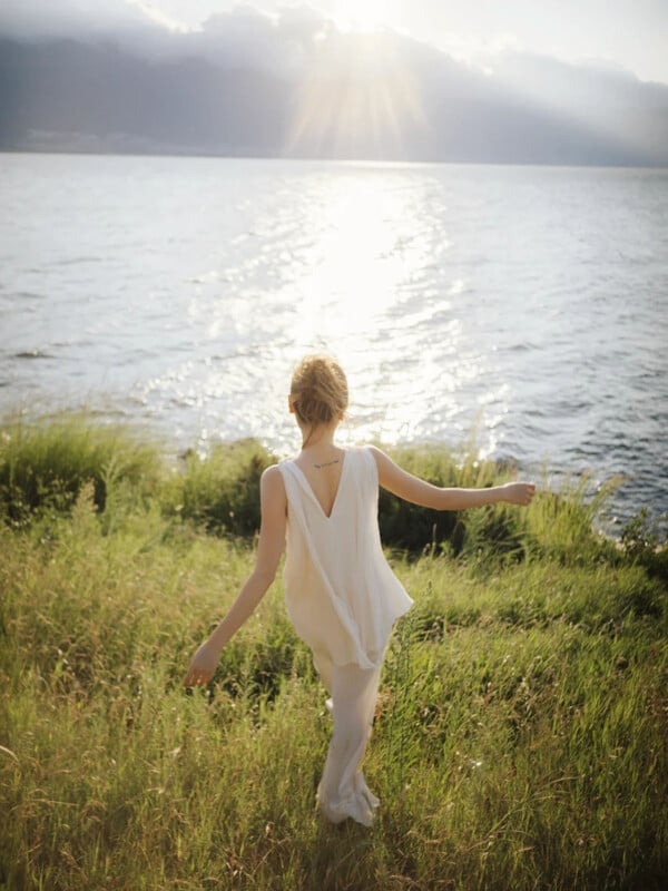 A woman in a flowing white dress walks through tall grass toward a calm lake, with sunlight reflecting off the water and distant mountains in the background.
